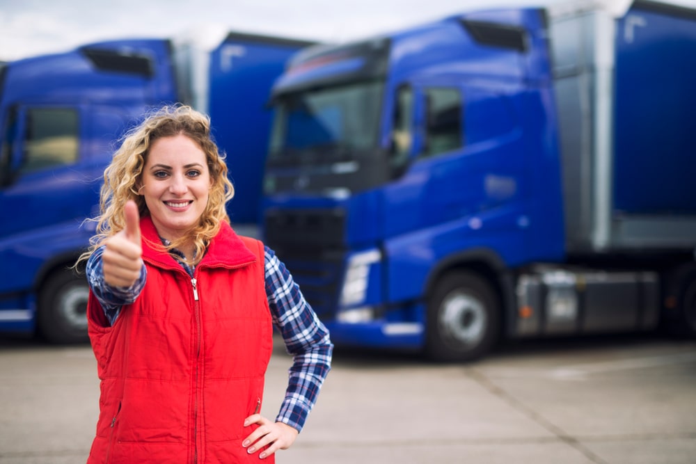 A girl in a red vest gives a thumbs-up in front of two blue trucks